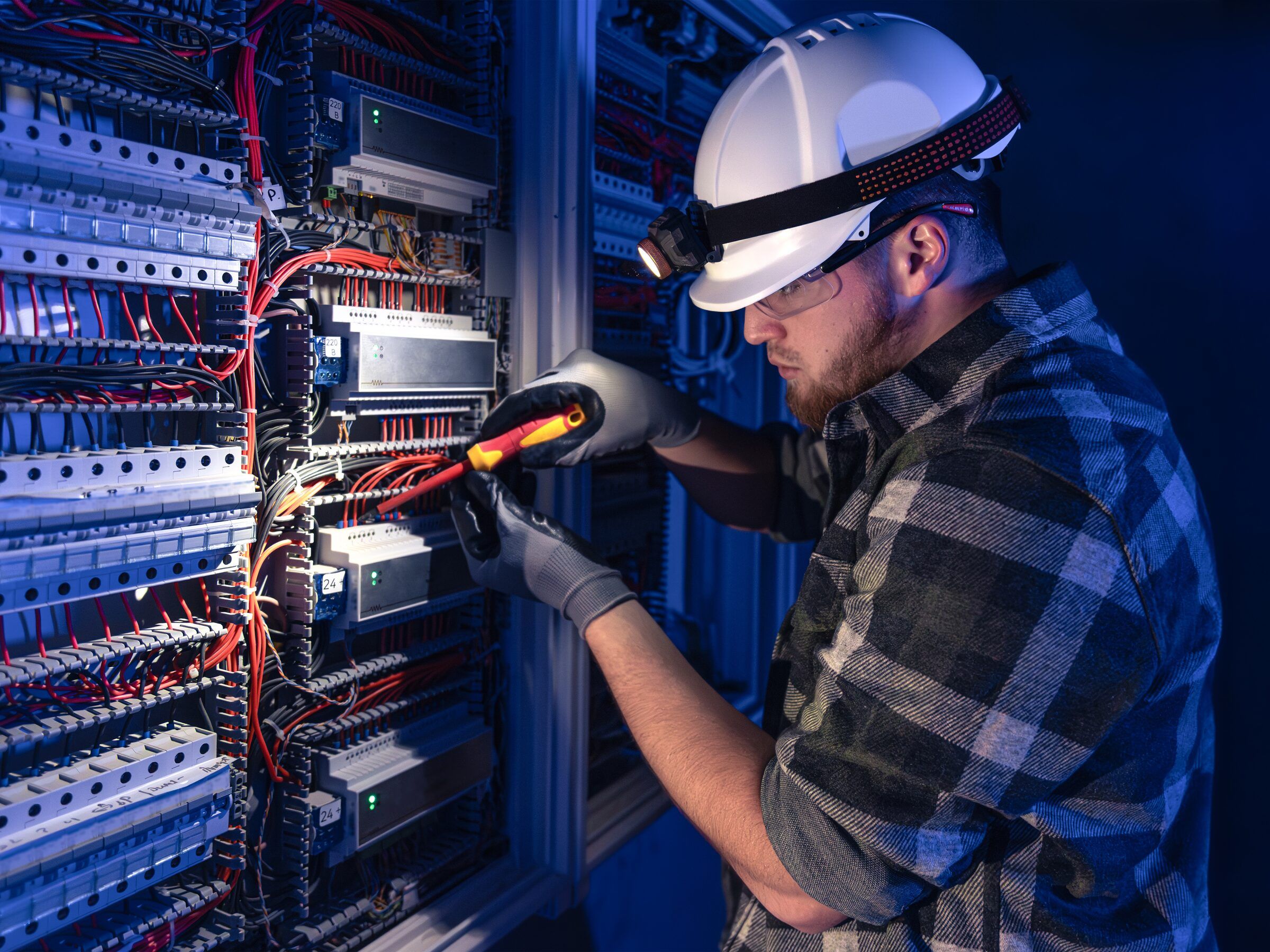Electrician in a hard hat and head torch working on a control panel with an insulated screwdriver